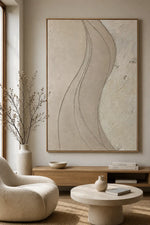 Large framed abstract wall art with flowing wavy lines in warm beige and cream tones, displayed in a modern living room above a wooden console with a cream chair and round coffee table.