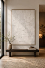 A large vertical framed wall art piece with a textured off-white surface, displayed in a minimalist interior setting above a dark wooden bench with a vase and books.