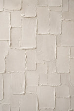 A close-up of a textured wall or panel with irregular rectangular sections in warm off-white and cream tones, showing raised plaster-like surfaces and subtle shadowing.