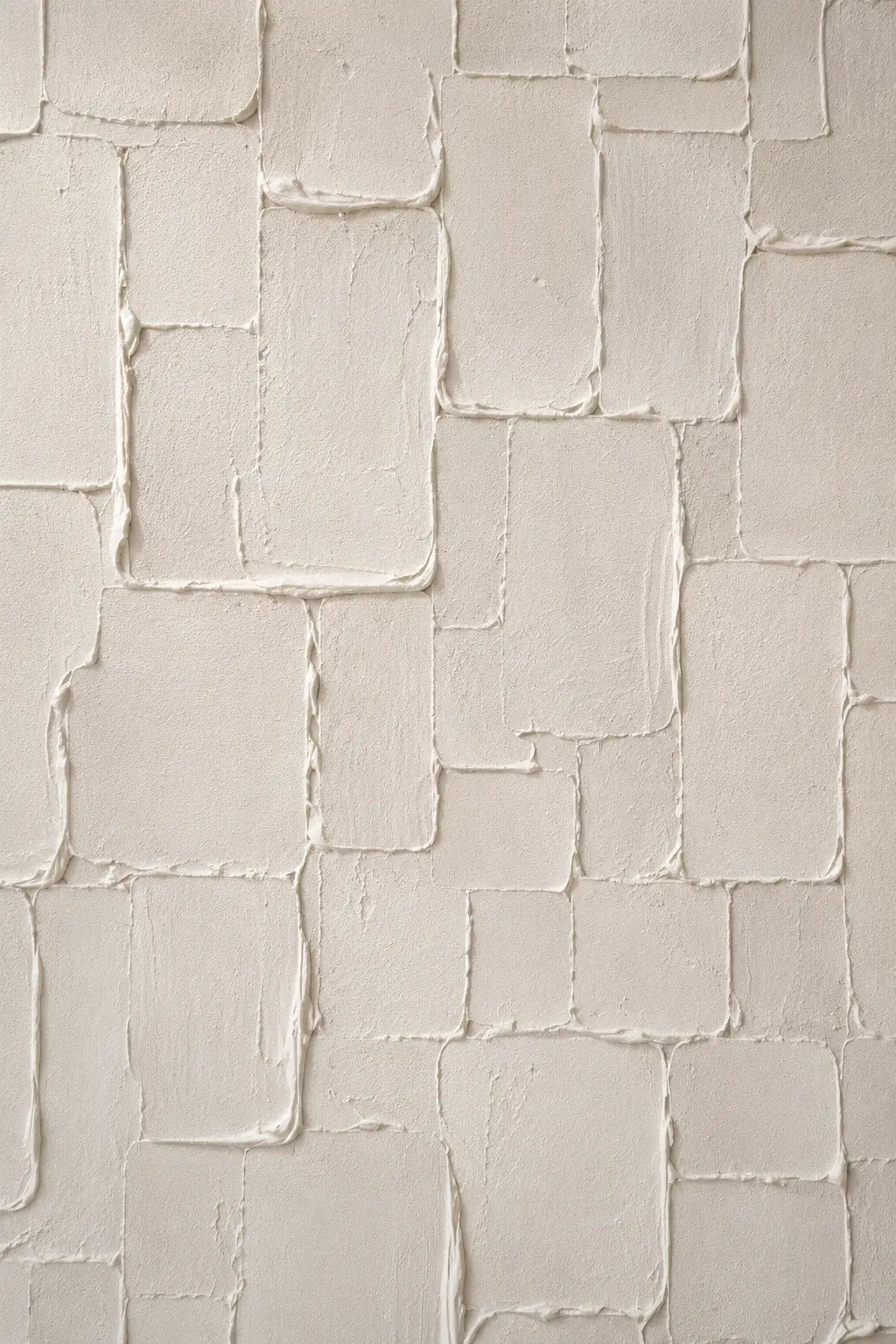 A close-up of a textured wall or panel with irregular rectangular sections in warm off-white and cream tones, showing raised plaster-like surfaces and subtle shadowing.