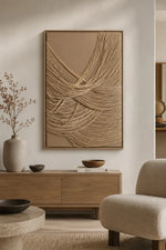 A large framed abstract wall art piece with layered beige and tan cord-like lines hangs above a wooden console in a styled living room with a chair, vase, and bowls.