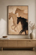 A framed wall art print showing three horses in warm neutral tones, displayed above a light wood console table in a styled interior with a vase and books.