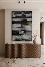 A large vertical abstract canvas print with textured black, gray, white, and subtle gold brushstrokes, displayed on a beige wall above a modern wavy wooden console table in a styled interior.