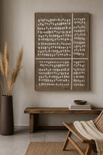A large four-panel abstract wall art piece with white organic dot patterns on a taupe-brown background, displayed in a styled room above a wooden bench with books and a chair.