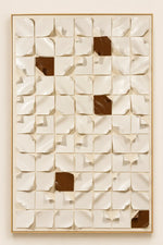 A framed abstract wall art piece featuring a grid of 3D curled paper-like panels in warm off-white tones with a few dark brown square accents, mounted against a matching light beige wall.