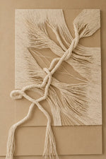 A square beige wall-hanging or textile art piece with layered fringed strands and a central knotted rope detail, displayed against a matching beige background.