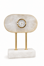 A modern table clock featuring a polished cream-colored onyx stone top with a gold-tone rim and a central white clock face, supported by two slim gold brass legs on a rectangular white marble base, isolated on a white background.