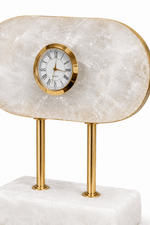 A modern tabletop clock with a polished gold-tone metal frame and two matching gold-tone cylindrical legs, set against a white marble clock face and a larger white marble base. The brand name 'GUST' is visible on the clock dial.