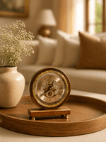 A vintage-style wooden mantel clock with a brass-toned frame and Roman numerals sits on a round wooden tray in a softly lit living room setting.