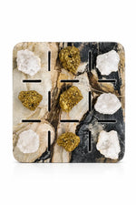 A square decorative tray or coaster with a faux-marble pattern in beige, black, and brown, featuring nine cutout slots and six rough crystal-like stones placed inside.