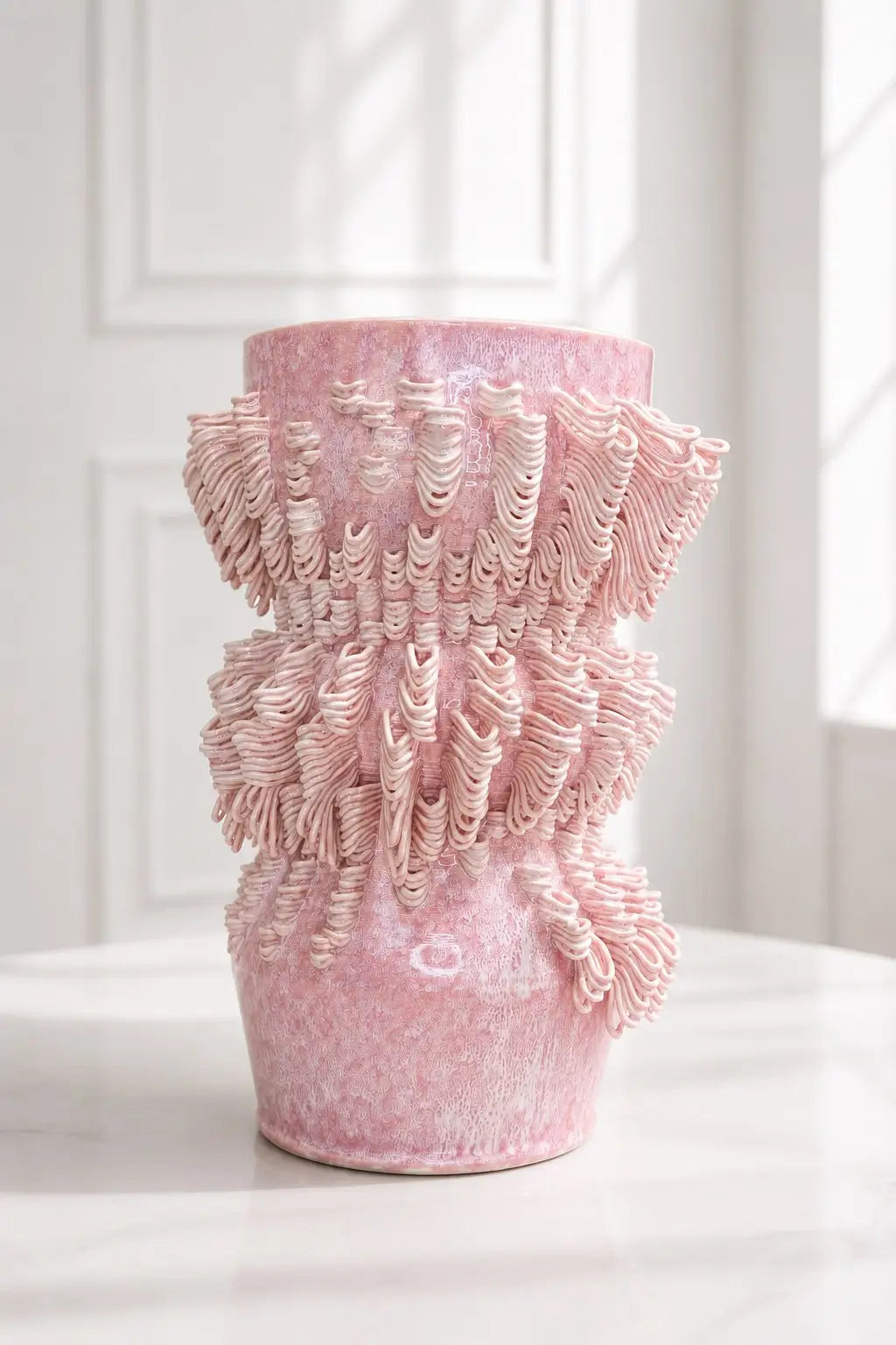 A pink textured ceramic vase with a flared, ruffled top and a tapered base, displayed on a white surface against a white paneled wall.
