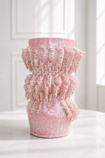 A pink textured ceramic vase with a flared, ruffled top and a tapered base, displayed on a white surface against a white paneled wall.