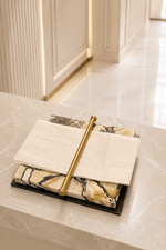 A luxury stationery set featuring a black tray with stacked marble notepads in cream, beige, and black, adorned with a gold pen and a folded cream paper napkin, set on a polished marble counter.