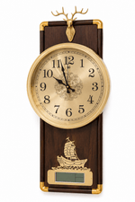 A decorative wall clock with a dark wood rectangular body, gold-tone metal trim, a round cream clock face with black numerals, a deer head topper, and a small ship illustration near a digital display window, shown on a white background.