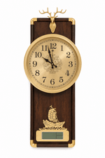 A vertical wooden wall clock with a dark brown wood body, gold-tone metal trim, a round cream clock face with black numerals, a deer head topper, and a ship engraving near the bottom.