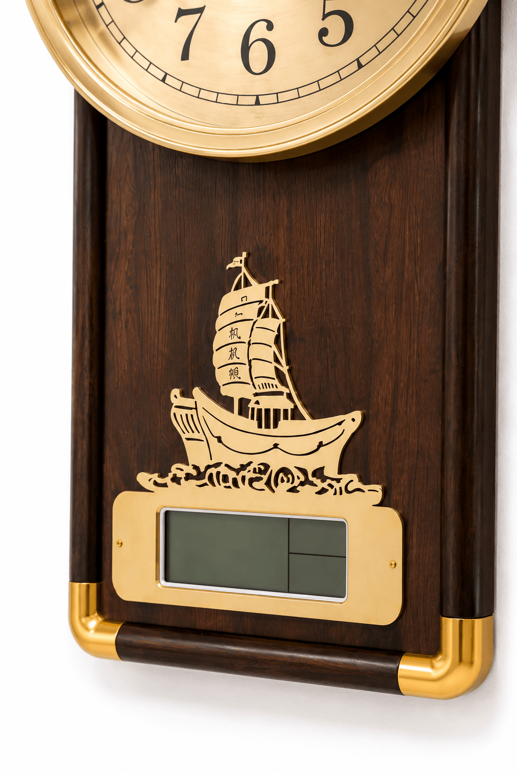 A close-up of a wall clock with a dark wood body, gold-tone brass bezel and trim, and an engraved sailing ship motif above a digital display window.