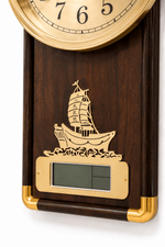 A close-up of a wall clock with a dark wood body, gold-tone brass bezel and trim, and an engraved sailing ship motif above a digital display window.