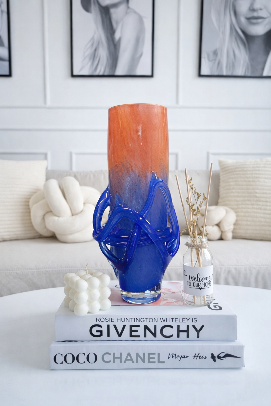 Blue and orange vase on books with a white couch and framed pictures in the background