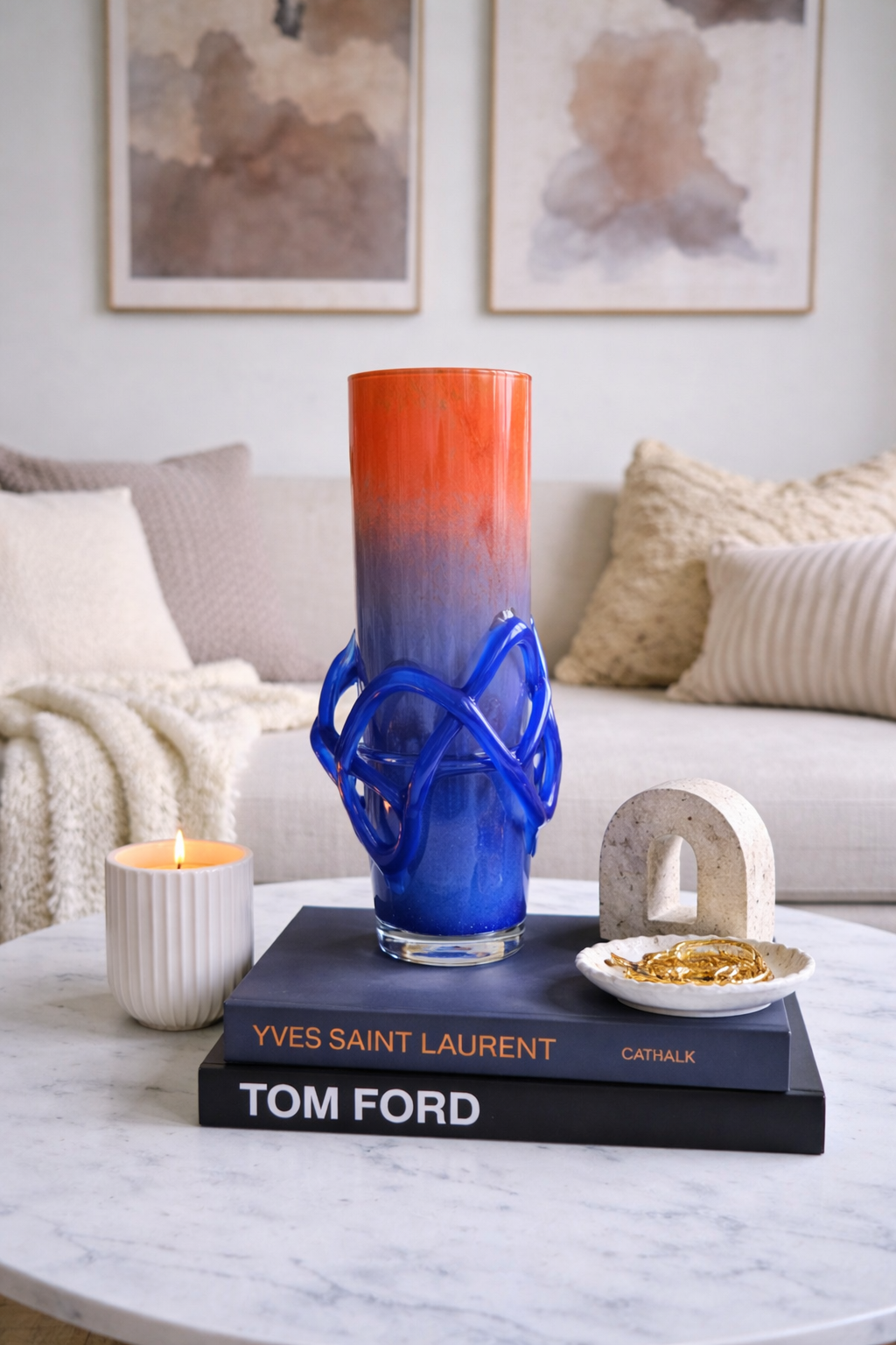 Blue and red glass vase on books with a candle and decorative items on a marble table.