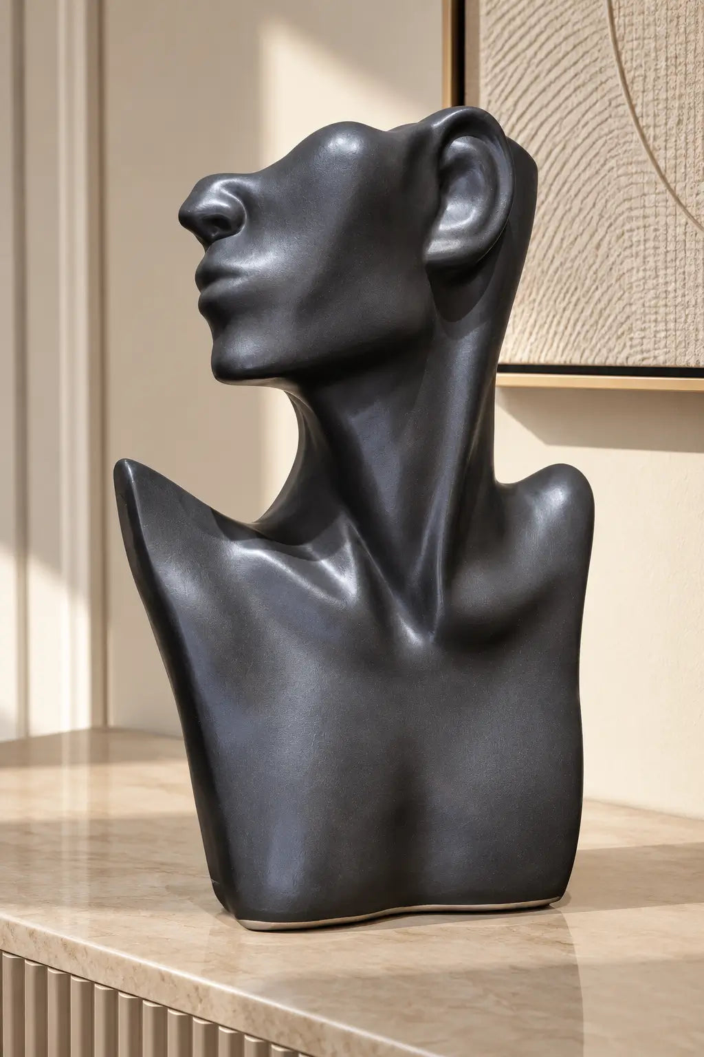 A matte black abstract sculptural bust of a human head and shoulders, displayed on a light beige stone countertop in a modern interior setting.