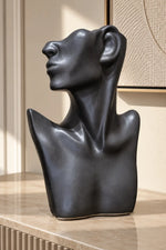 A matte black abstract sculptural bust of a human head and shoulders, displayed on a light beige stone countertop in a modern interior setting.
