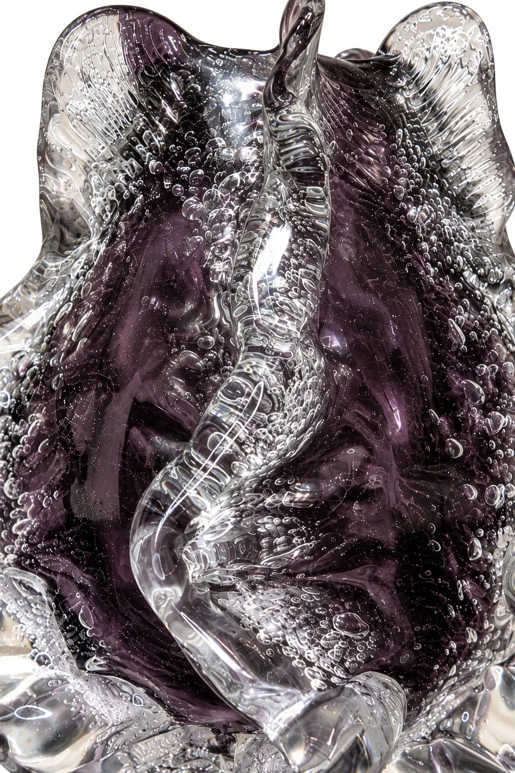 A close-up of a sculptural glass object with a wavy, organic form, featuring a translucent clear outer layer and deep purple inner areas filled with bubble details, set against a plain white background.