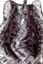 A close-up of a sculptural glass object with a wavy, organic form, featuring a translucent clear outer layer and deep purple inner areas filled with bubble details, set against a plain white background.
