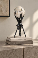 A decorative globe sculpture with a black tripod-style stand displayed on top of two books on a stone surface in a styled interior setting.