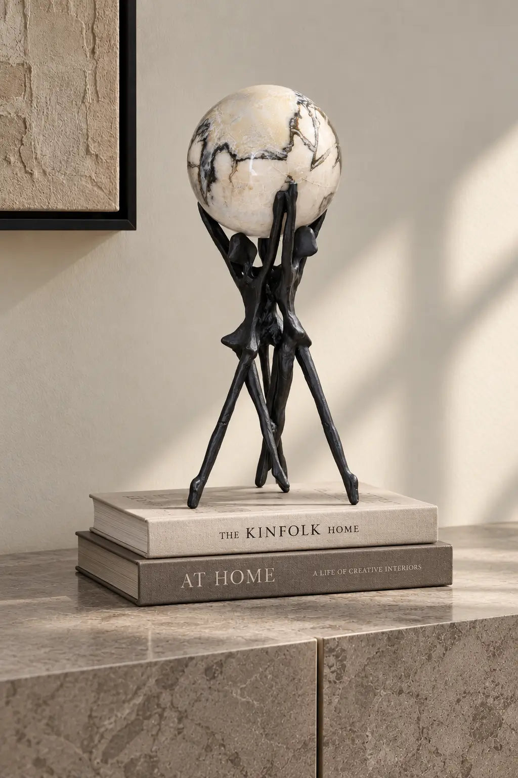 A decorative globe sculpture with a black tripod-style stand displayed on top of two books on a stone surface in a styled interior setting.