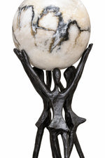 A decorative sculpture featuring a large spherical stone orb with cream, white, and dark gray marbling, supported by a dark metallic bronze or black metal stand shaped like abstract human figures.