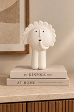 A white sculptural elephant figurine with a sunburst mane stands on top of two stacked books on a beige marble surface in an interior setting.