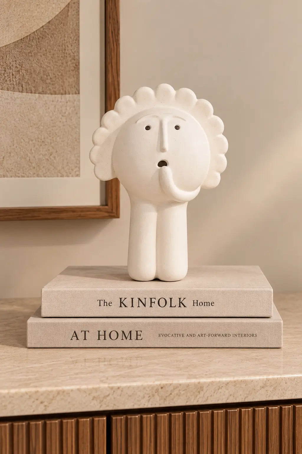 A white sculptural elephant figurine with a sunburst mane stands on top of two stacked books on a beige marble surface in an interior setting.