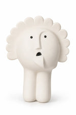 A minimalist, sculptural ceramic figurine of a face with a sunburst or flower-like crown, standing on two legs. The figure is off-white with small black eyes and a black mouth, set against a clean white background.