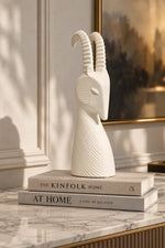 A white sculptural goat head decor piece with curved horns, displayed on top of two stacked books on a marble surface in an elegant interior setting.
