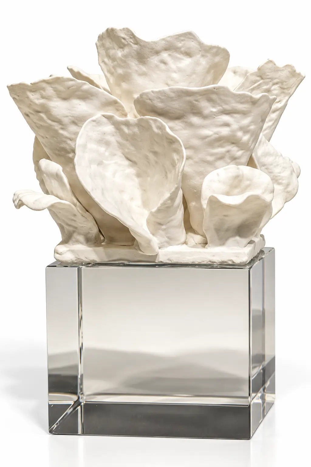 A white sculptural floral arrangement with textured, organic petals displayed on a clear square glass pedestal against a white background.