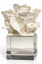 A white sculptural floral arrangement with textured, organic petals displayed on a clear square glass pedestal against a white background.