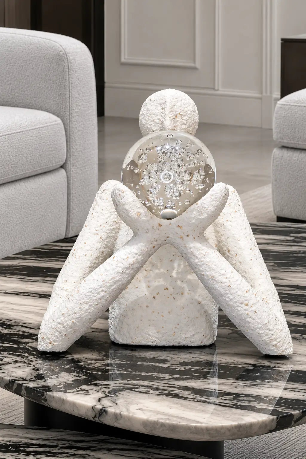 A decorative white textured figurine with a clear glass sphere head containing a snowflake design, displayed on a black and white marble coffee table in a modern living room setting.