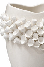 A close-up of a white ceramic vase featuring a textured, organic shape adorned with a dense cluster of sculpted, 3D flower buds around the upper portion.