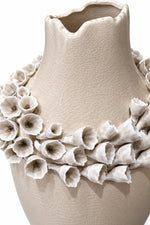 A beige ceramic vase with a textured, crackled surface, decorated with a wide collar of white and beige sculpted flower-like ruffles around the neck, shown against a plain white background.
