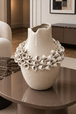 A large textured ceramic vase in a warm cream-beige color, decorated with a wide band of sculpted white calla lilies around the middle, displayed on a modern round coffee table in a stylishly decorated living room.