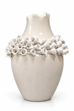 A cream-colored ceramic vase with a textured finish, featuring a wide collar decorated with a ring of sculpted white flowers, set against a plain white background.