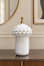 A white ceramic decorative lidded jar with a geometric textured dome lid and a small gold finial, displayed on a wooden console table in an elegant interior setting.