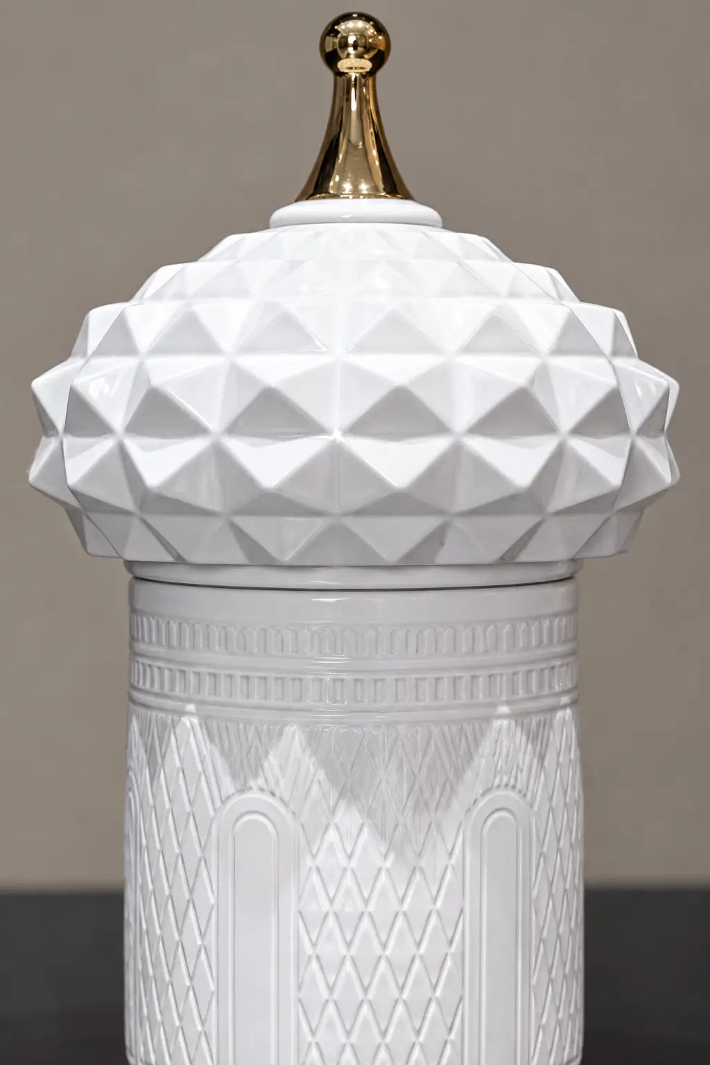 A white ceramic lidded jar with a faceted geometric dome lid and a polished gold finial on top, set against a neutral gray background.