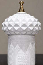 A white ceramic lidded jar with a faceted geometric dome lid and a polished gold finial on top, set against a neutral gray background.