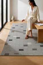 A woman in cream pajamas sits on a wooden bench beside a large gray area rug with a grid pattern of square blocks in gray, charcoal, and beige tones, set in a bright room with light wood floors.