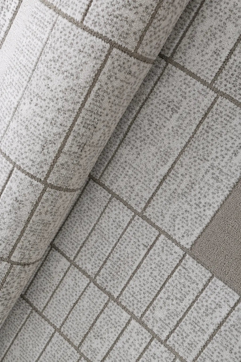 A close-up of a textured area rug with a light gray and white speckled pattern, featuring a grid design and a solid taupe-gray accent block.