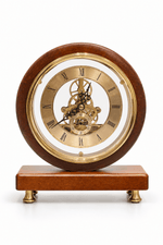 A round wooden mantel clock with a polished brown wood frame, a gold-toned brass inner ring, and a visible mechanical gear train through the center. The clock sits on a matching rectangular wooden base with small brass feet, photographed against a clean white background.