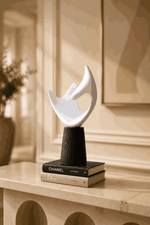 A modern abstract white sculpture on a black cylindrical pedestal, styled on top of two designer books (Chanel and Dior) on a beige stone mantelpiece in a warm, elegant interior.