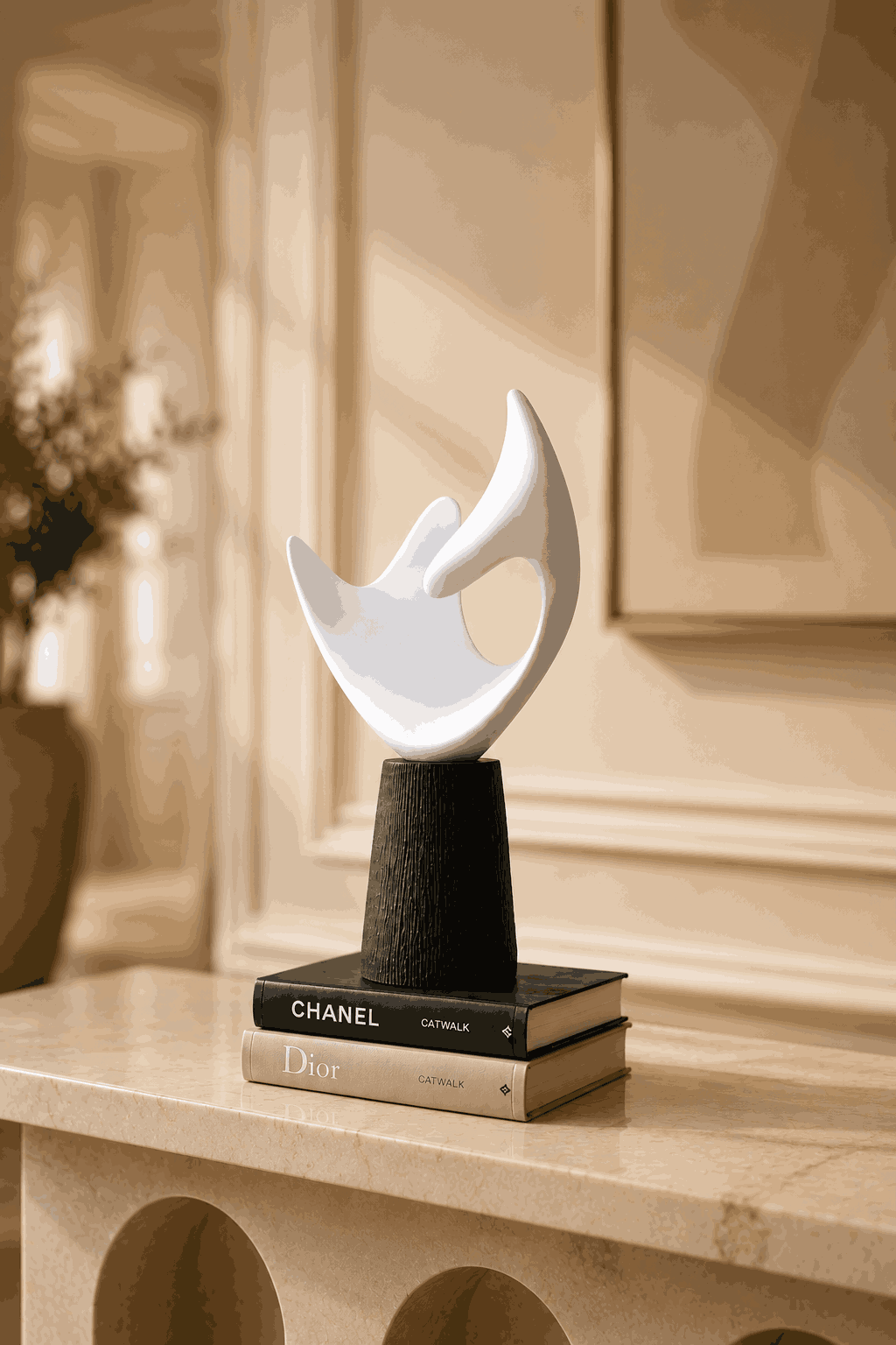 A modern abstract white sculpture on a black cylindrical pedestal, styled on top of two designer books (Chanel and Dior) on a beige stone mantelpiece in a warm, elegant interior.