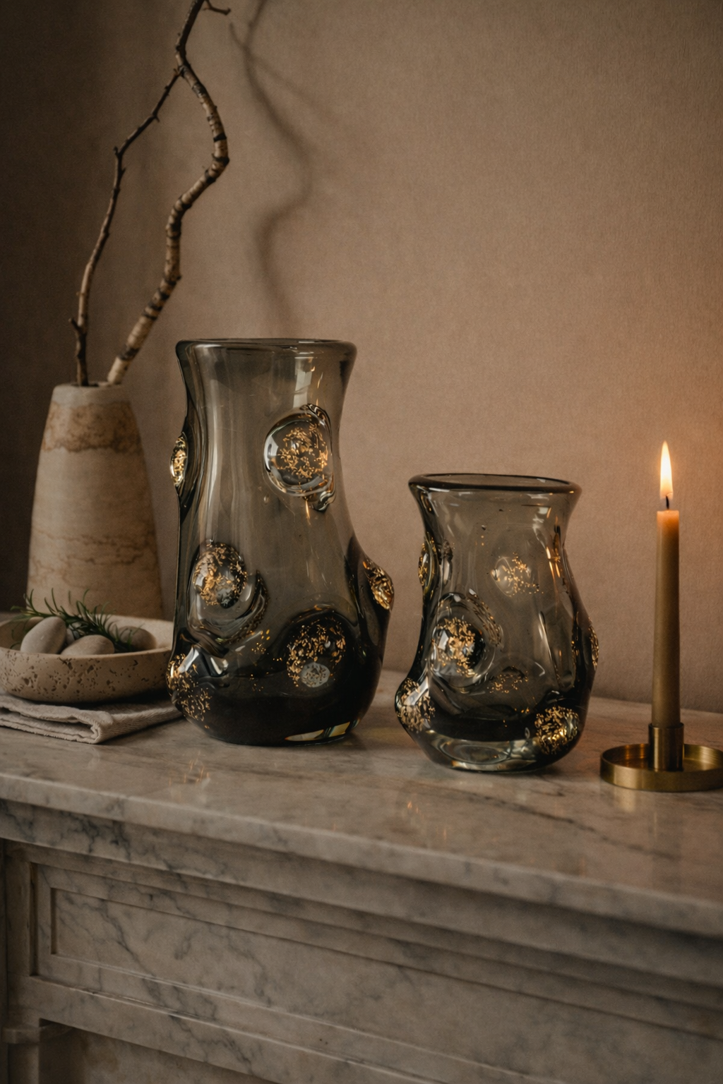 Two glass vases with decorative elements on a marble surface with a candle in the background.